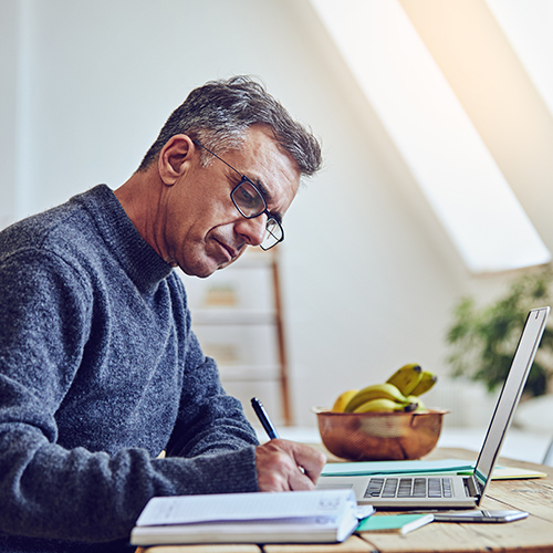 older male taking notes on desk with laptop and notebooks on desk